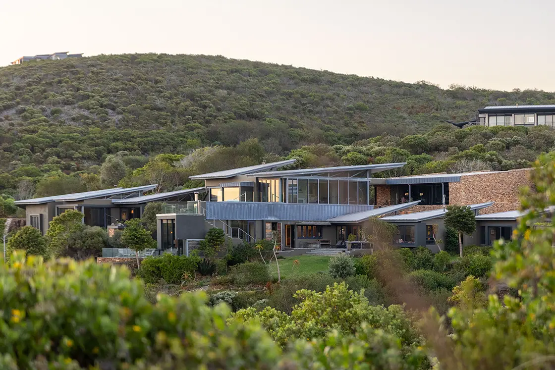 Perfect Hideaways real estate property: Modern house with glass and stone façade nestled in lush greenery. Rolling hills in the background under a soft, cloudy sky, conveys tranquility. Knysna, Ocean’s Edge.