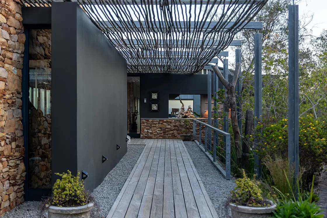 Perfect Hideaways real estate property: Wooden walkway leading to a modern house with stone and black walls, covered by a slatted pergola. Potted plants and trees line the path, creating a serene atmosphere. Knysna, Ocean’s Edge.