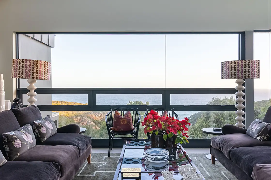 Perfect Hideaways real estate property: Modern living room with plush sofas, patterned cushions, and bold lamps. Large window displays scenic ocean and coastal view, creating a serene atmosphere. Knysna, Ocean’s Edge.