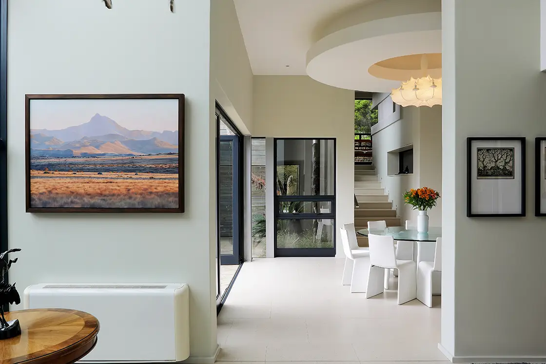Perfect Hideaways real estate property: Modern dining area with white chairs around a glass table. Landscape painting on a beige wall, warm natural light, and a circular ceiling light enhance the calm ambiance. Knysna, Ocean’s Edge.