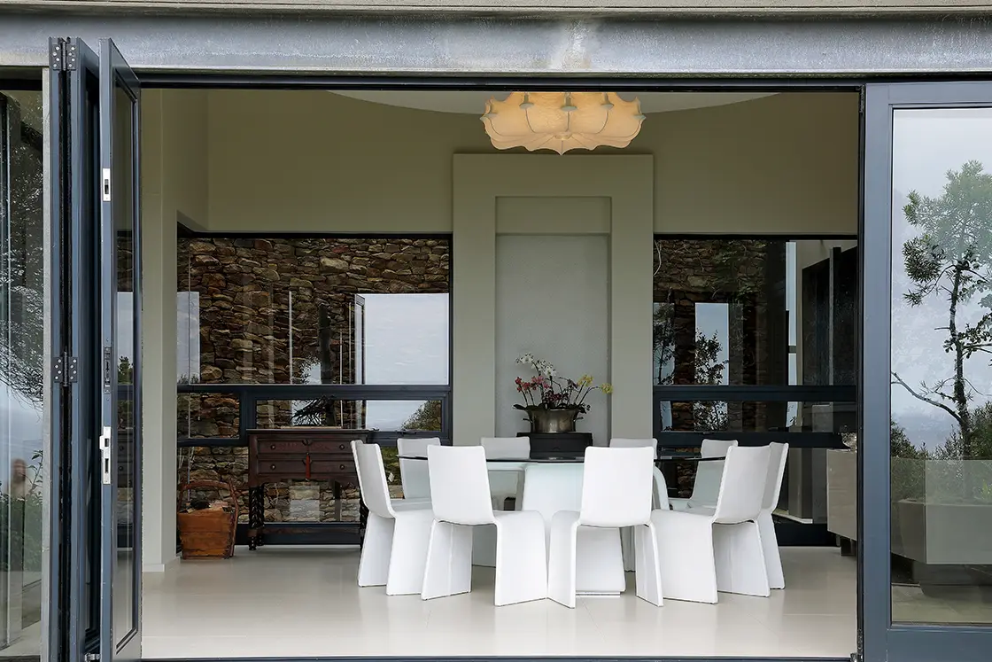 Perfect Hideaways real estate property: Modern dining room with glass doors open to a scenic view. White table and chairs, stone walls, potted plants, and soft lighting create an inviting atmosphere. Knysna, Ocean’s Edge.