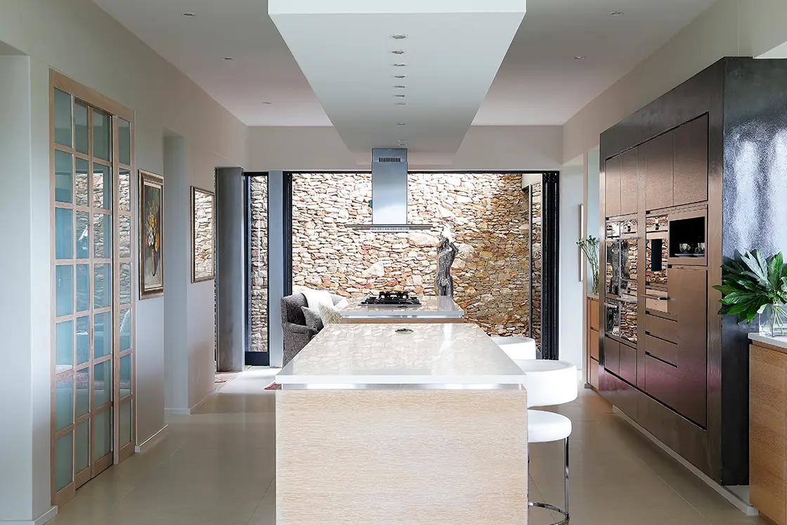 Perfect Hideaways real estate property: Modern kitchen with sleek wooden cabinetry, a long island with bar stools, and a stainless-steel range hood. Stone accent wall offers a rustic touch. Knysna, Ocean’s Edge.