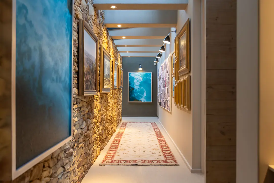 Perfect Hideaways real estate property: A narrow hallway with rustic stone and framed art on the walls, lit by overhead lights. A patterned rug runs along the floor, creating an inviting atmosphere. Knysna, Ocean’s Edge.