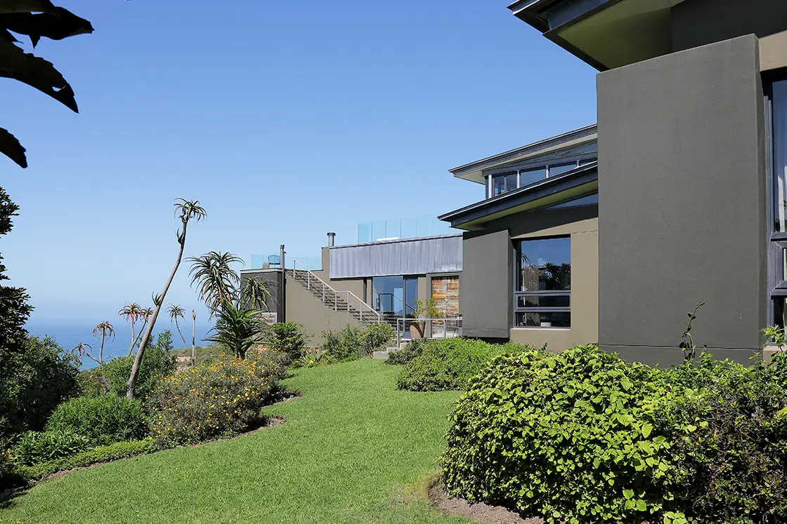 Perfect Hideaways real estate property: Modern house with large windows in a lush garden setting, overlooking the ocean under a clear blue sky. The scene is tranquil and spacious. Knysna, Ocean’s Edge.