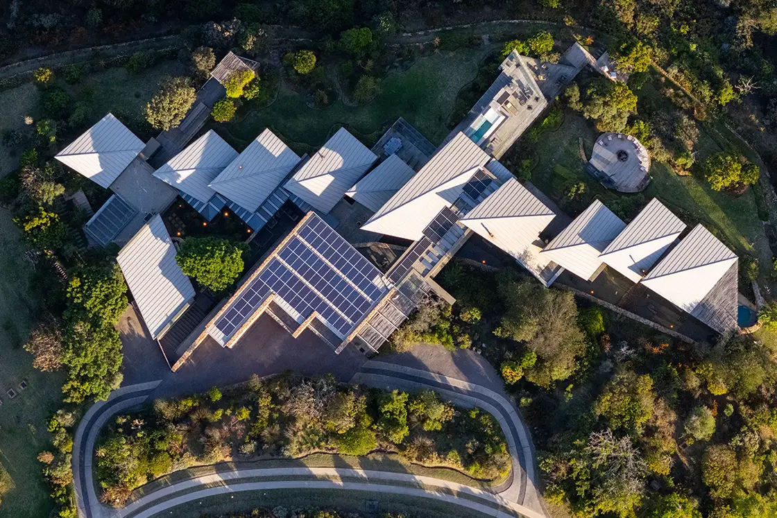 Perfect Hideaways real estate property: Aerial view of a large, modern building with a zigzag roof and solar panels, surrounded by lush greenery and winding roads, under soft light. Knysna, Ocean’s Edge.