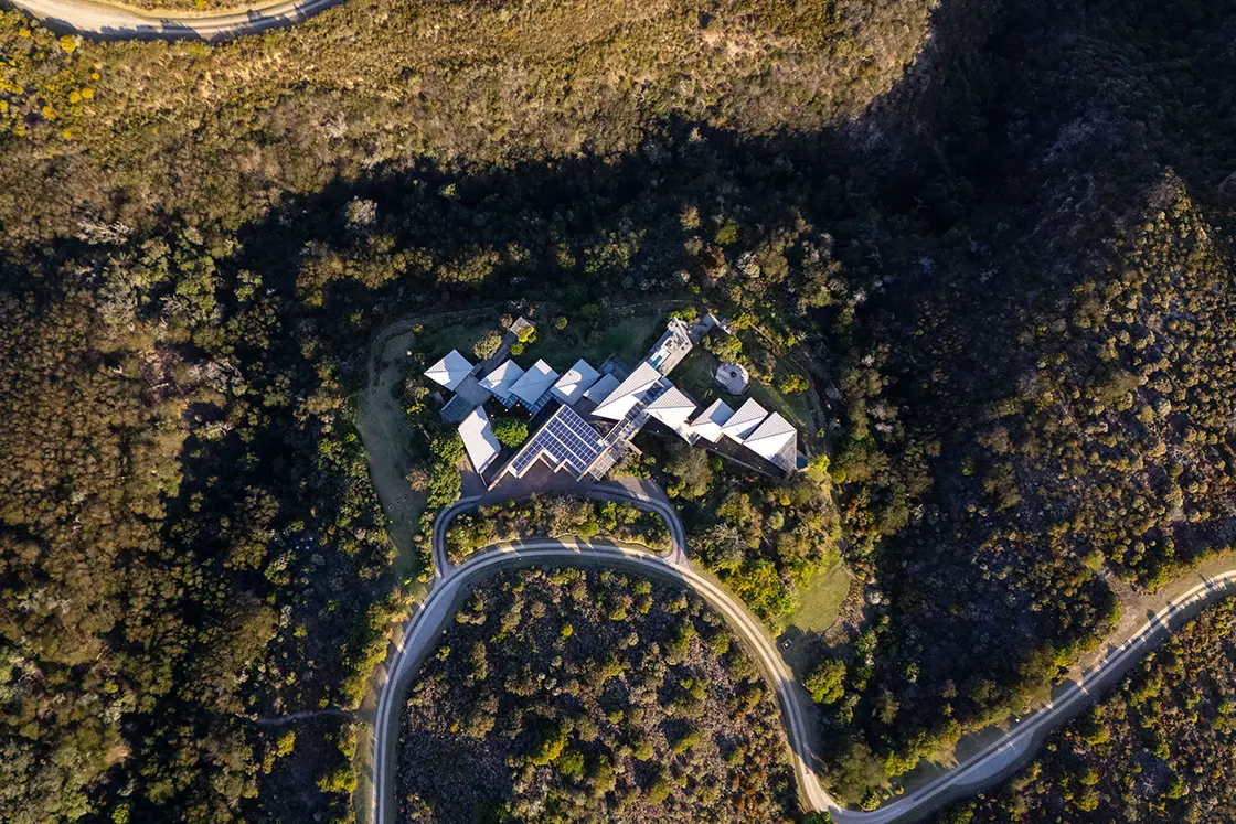 Perfect Hideaways real estate property: Aerial view of a large, modern house with a distinct geometric design, surrounded by dense, lush greenery and winding roads in a mountainous area. Knysna, Ocean’s Edge.