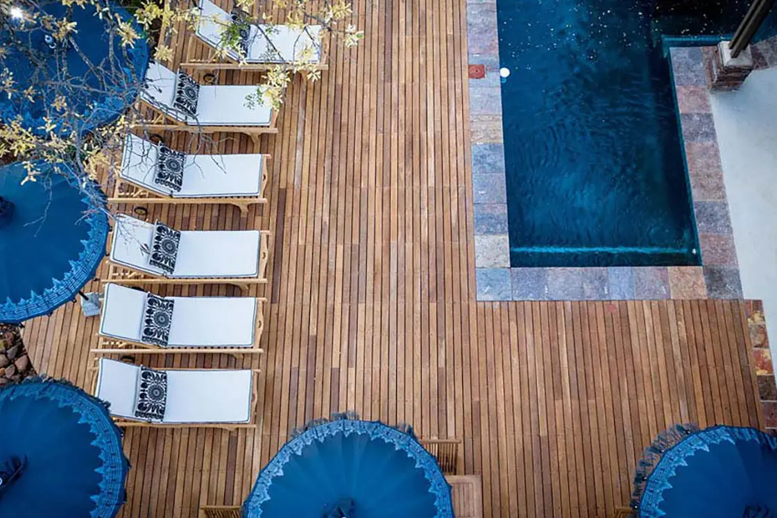 Perfect Hideaways real estate property: Overhead view of a wooden deck featuring six white lounge chairs with patterned cushions beside blue parasols, adjacent to a small rectangular pool. Relaxing ambiance. Hoedspruit Wildlife Estate, Safari Moon Luxury Bush Lodge.