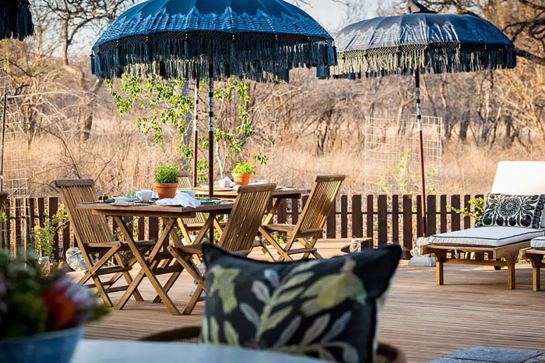 Perfect Hideaways real estate property: Outdoor deck with a wooden table and chairs under blue fringed umbrellas. Sun loungers with patterned cushions nearby. Serene, rustic setting. Hoedspruit Wildlife Estate, Safari Moon Luxury Bush Lodge.