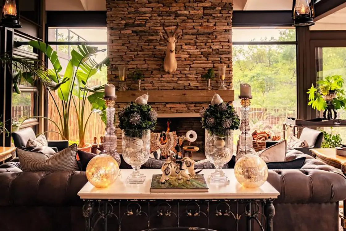 Perfect Hideaways real estate property: Cozy living room with rustic stone fireplace, deer head, large plants, and rich leather seating. Warm, inviting ambiance with elegant, eclectic decor. Hoedspruit Wildlife Estate, Safari Moon Luxury Bush Lodge.