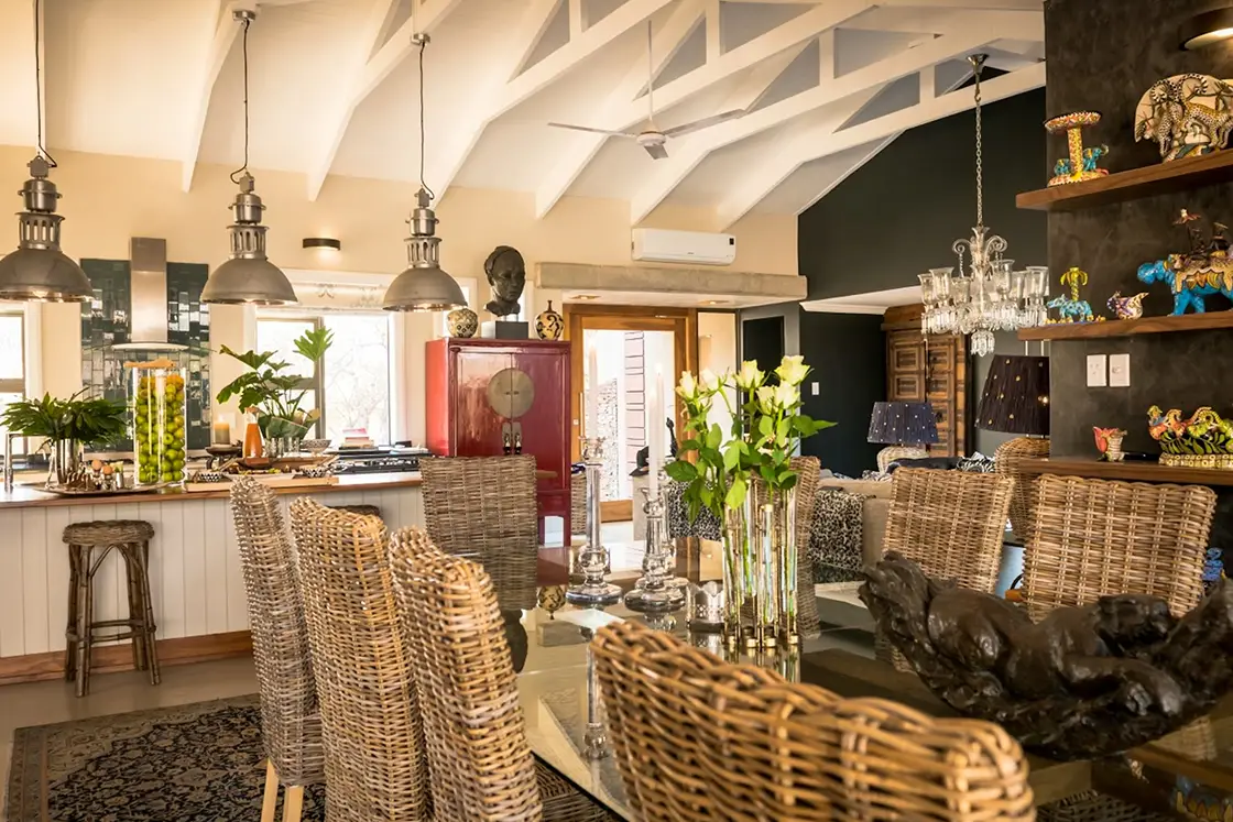 Perfect Hideaways real estate property: Spacious dining area with woven chairs surrounds a glass table adorned with fresh flowers. Industrial lights hang above. Cozy, eclectic decor with warm tones. Hoedspruit Wildlife Estate, Safari Moon Luxury Bush Lodge.