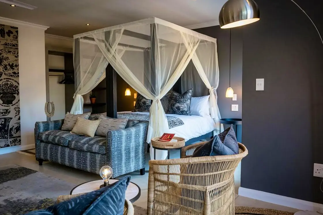 Perfect Hideaways real estate property: Elegant bedroom with a four-poster bed draped in sheer curtains, cozy sofa, and rattan chair. Warm lighting and modern, serene ambiance. Hoedspruit Wildlife Estate, Safari Moon Luxury Bush Lodge.