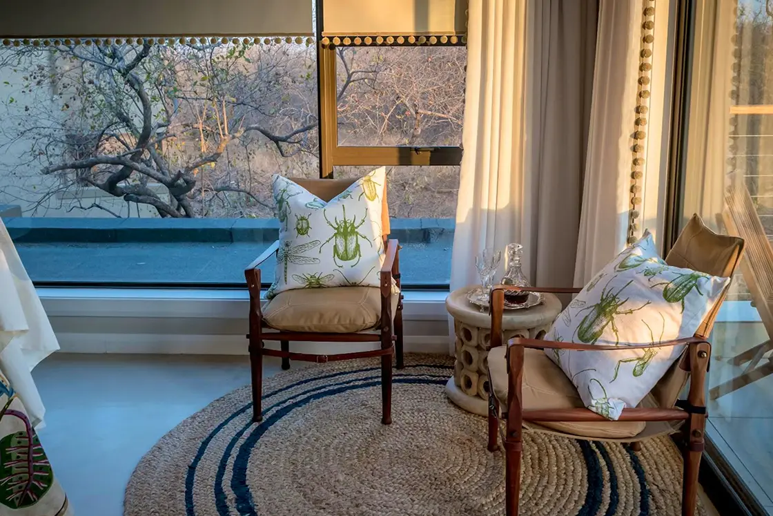 Perfect Hideaways real estate property: Cozy sunlit corner with two wooden chairs and insect-patterned cushions, small table with glassware, round rug, large window, and tree views outside. Hoedspruit Wildlife Estate, Safari Moon Luxury Bush Lodge.