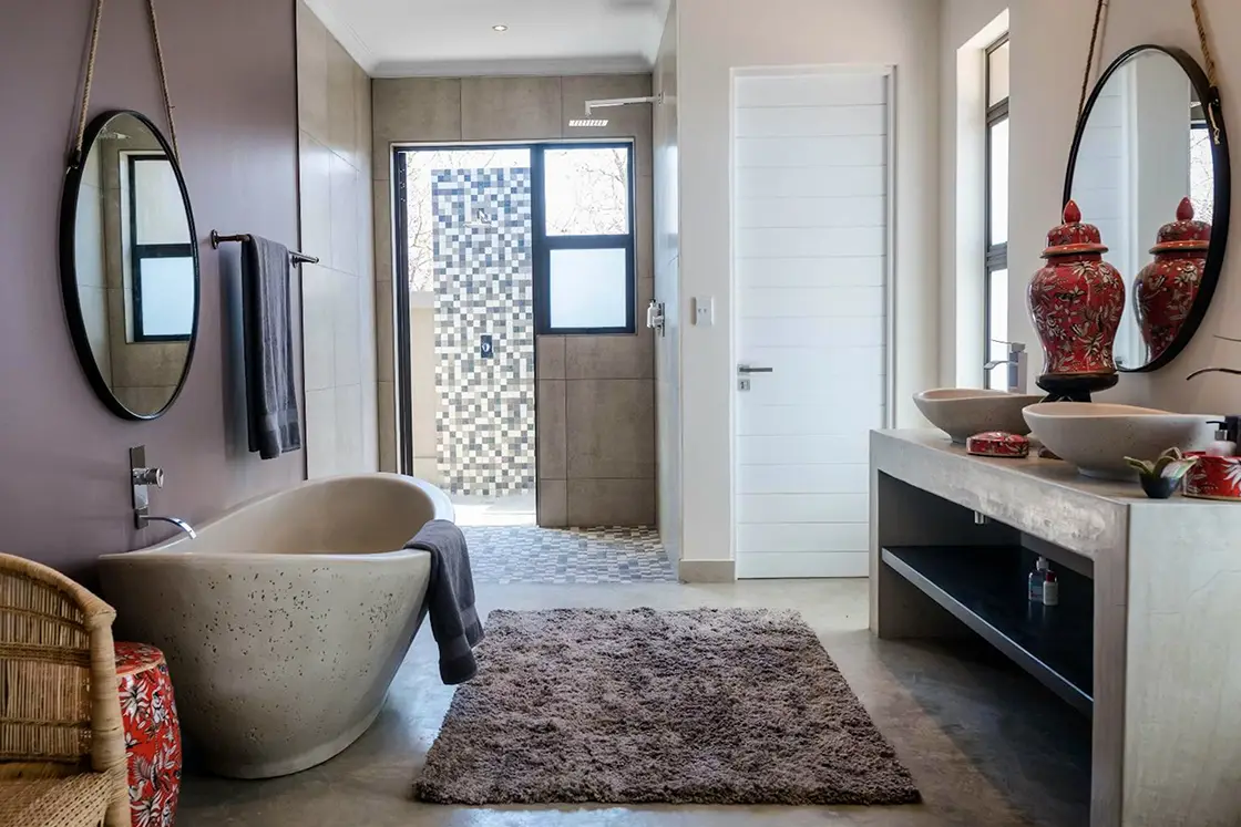 Perfect Hideaways real estate property: Spacious bathroom with a freestanding oval bathtub, round mirrors, twin red vases on the counter, soft rug, and large tile shower, evoking elegance. Hoedspruit Wildlife Estate, Safari Moon Luxury Bush Lodge.