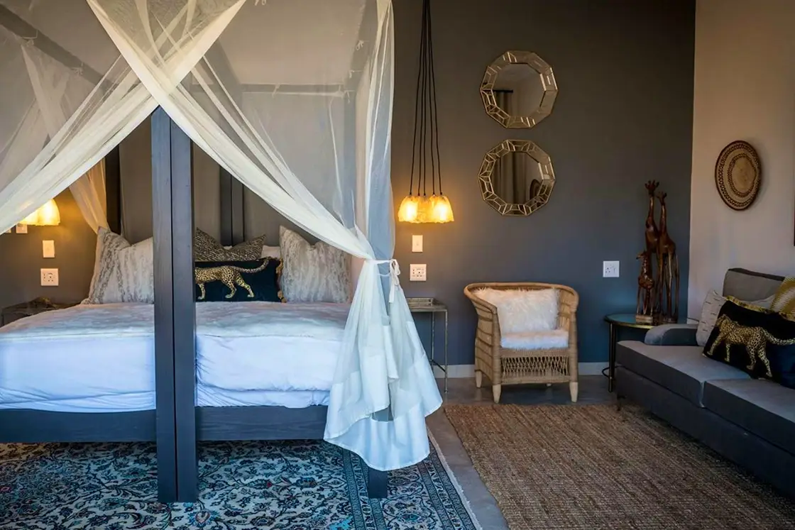 Perfect Hideaways real estate property: Stylish bedroom with a canopy bed draped in white fabric, decorative mirrors on a dark wall, woven chair, and ambient lighting creating a cozy atmosphere. Hoedspruit Wildlife Estate, Safari Moon Luxury Bush Lodge.