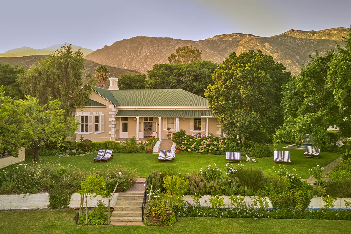 Perfect Hideaways real estate property: Elegant Cape Dutch-style farmhouse with a green metal roof, wide front veranda, manicured lawns, striped sun loungers, and lush gardens set against dramatic mountain scenery at sunset, Montagu, Montagu Vines Guesthouse.