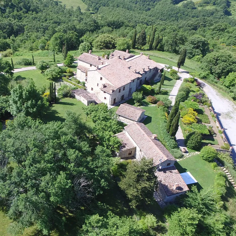 Perfect Hideaways real estate property: Aerial view of a large stone estate with multiple buildings, surrounded by trees and gardens, with a swimming pool and a driveway visible. Borgo Fastelli, Val d’Orcia.
