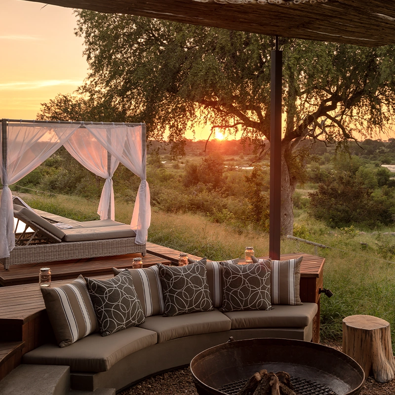 Perfect Hideaways real estate property: Outdoor lounge with a curved sofa and patterned cushions, facing a fire pit. A canopy bed nearby, set against a vibrant sunset over a lush landscape. N’Wambu Safari Lodge, Mjejane Private Game Reserve.