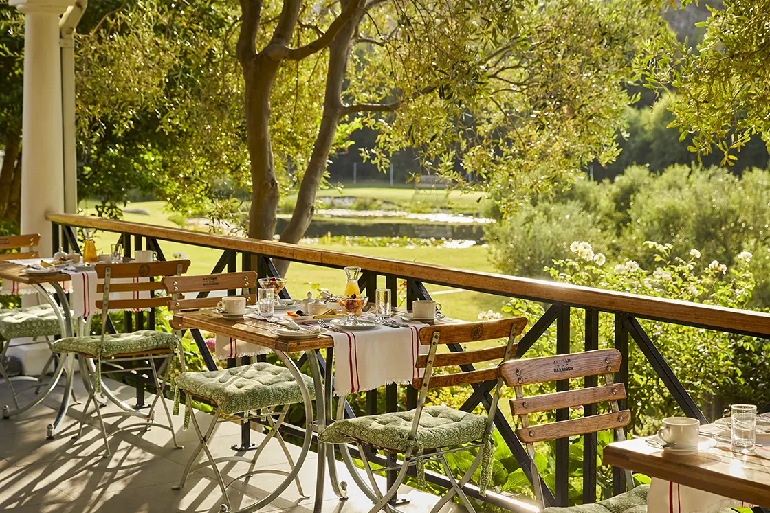 Perfect Hideaways real estate property: A picturesque outdoor dining area with wooden tables and chairs, set for breakfast. The setting is surrounded by lush greenery and a tranquil view of a field and trees in the background. Montagu, Montagu Vines Guesthouse.