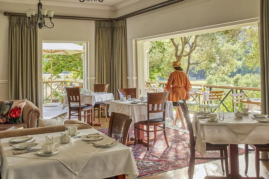 Perfect Hideaways real estate property: A cozy dining area with tables set for breakfast, featuring white tablecloths and dishes, large windows open to a view of greenery outside, and a person in casual attire walking towards the balcony. Montagu, Montagu Vines Guesthouse.