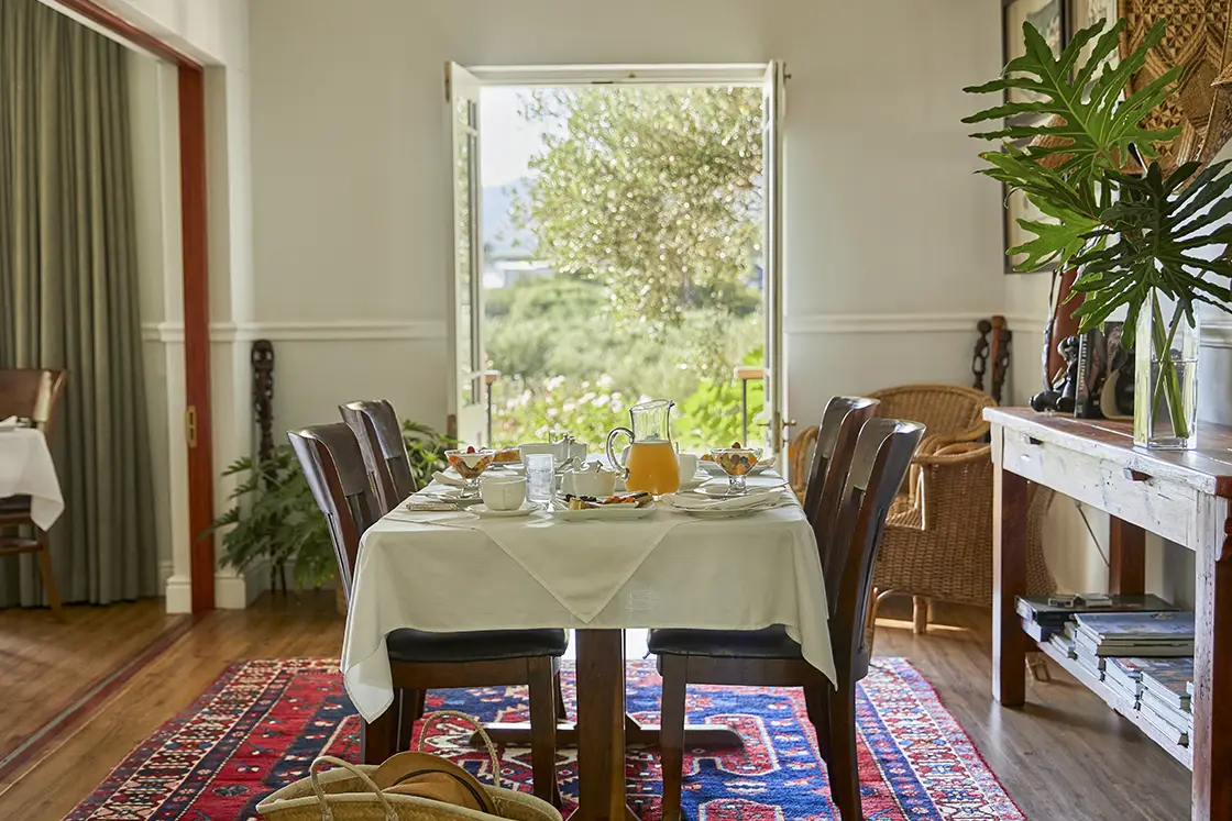 Perfect Hideaways real estate property: A dining table set with breakfast items, including a pitcher and glasses, in a room with open doors leading to a garden view. The floor is covered with a patterned rug, and there are chairs placed around the table. Montagu, Montagu Vines Guesthouse.