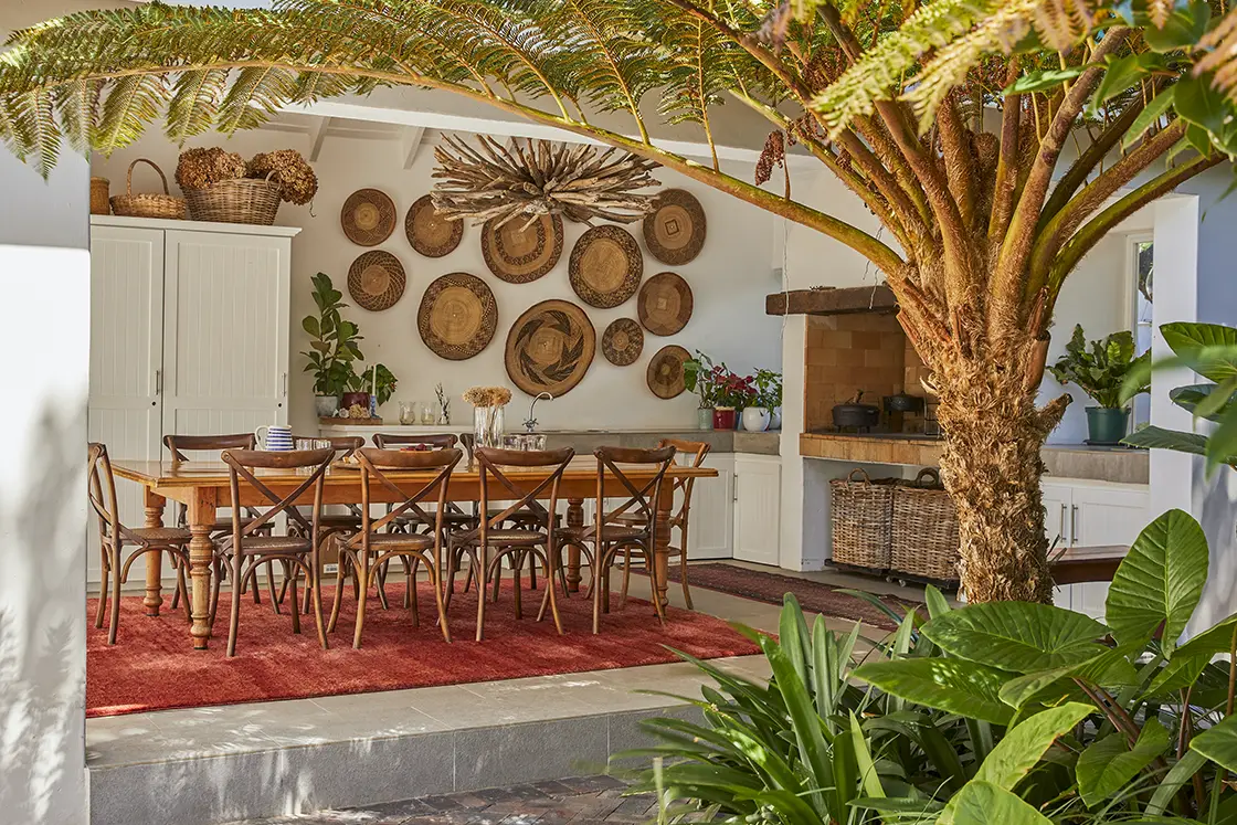 Perfect Hideaways real estate property: A dining area with a wooden table and chairs, surrounded by plants and decorated with woven baskets on the walls. A large tree is positioned in the corner, adding to the natural ambiance. Montagu, Montagu Vines Guesthouse.