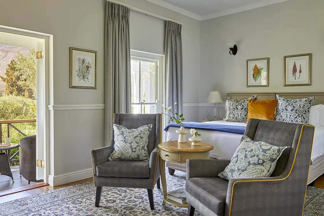 Perfect Hideaways real estate property: A cozy bedroom with two grey armchairs, a round wooden table between them, and a bed with decorative pillows. Natural light comes in through a window and door leading to an outdoor view. Soft gray curtains and framed artwork adorn the walls. Montagu, Montagu Vines Guesthouse.