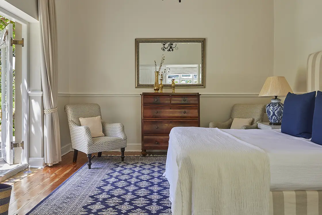 Perfect Hideaways real estate property: A well-lit bedroom featuring a bed with white linens, two blue pillows, and a decorative mirror above a wooden dresser. Two upholstered chairs are positioned near the window, and there is a patterned rug on the floor. Montagu, Montagu Vines Guesthouse.