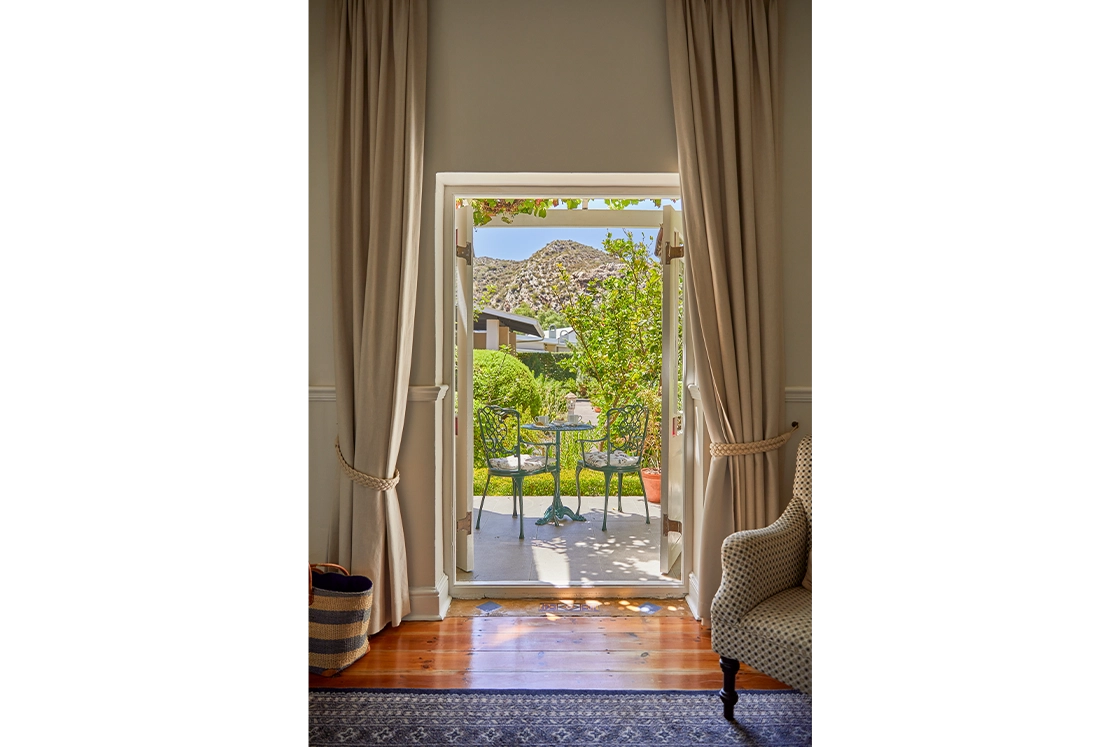 Perfect Hideaways real estate property: A view through an open doorway leading to a patio with a table and chairs surrounded by greenery, alongside a bathroom featuring a freestanding bathtub and framed pictures on the wall. Montagu, Montagu Vines Guesthouse.