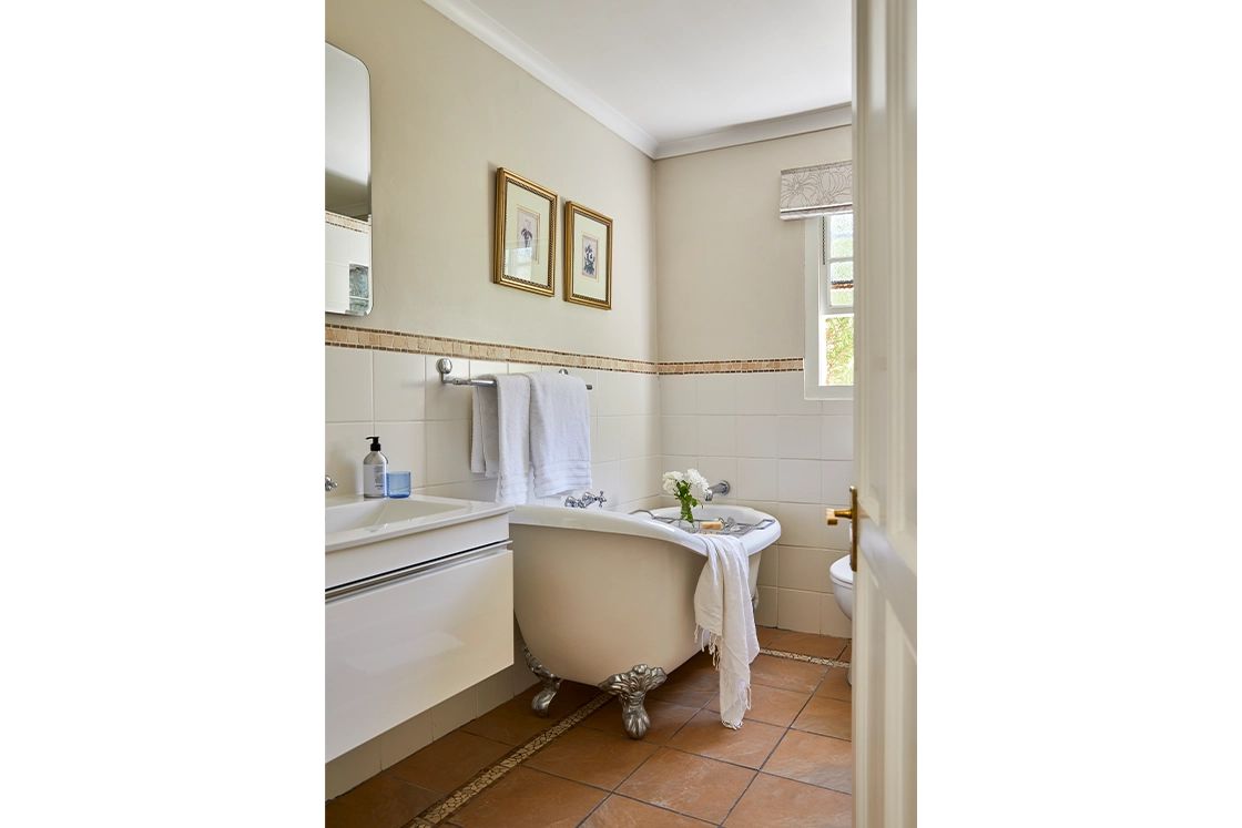 Perfect Hideaways real estate property: A view through an open doorway leading to a patio with a table and chairs surrounded by greenery, alongside a bathroom featuring a freestanding bathtub and framed pictures on the wall. Montagu, Montagu Vines Guesthouse.