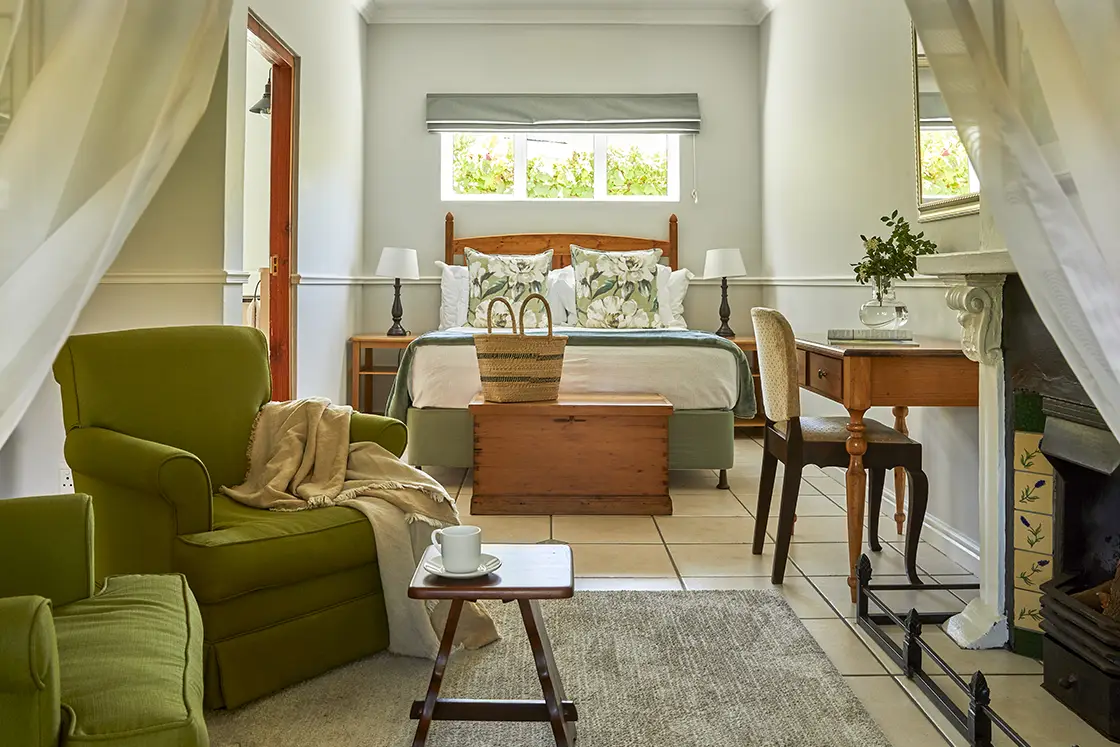 Perfect Hideaways real estate property: A cozy bedroom with a bed dressed in patterned linens, flanked by lamps. A green armchair and small coffee table are in the foreground, with a wooden desk and decorative items nearby. The room has a light and airy atmosphere with neutral tones. Montagu, Montagu Vines Guesthouse.