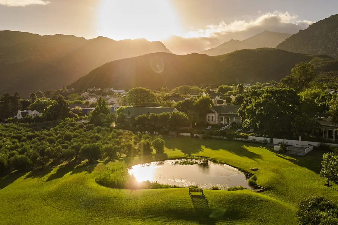 Perfect Hideaways real estate property: A scenic view of a landscape at sunset, featuring rolling hills, trees, a pond reflecting the sunlight, and buildings in the distance. Montagu, Montagu Vines Guesthouse.