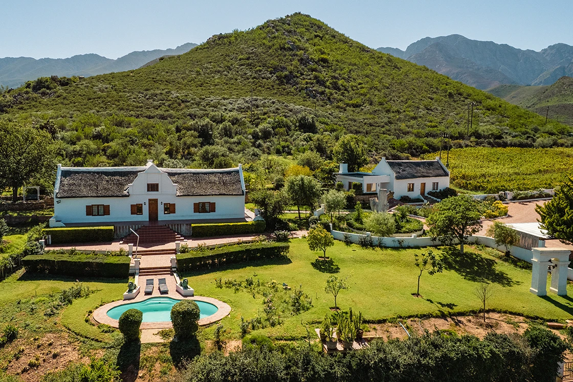Perfect Hideaways real estate property: Aerial view of a white farmhouse with a thatched roof, nestled in lush green hills. A well-manicured garden and oval pool are in the foreground. Scenic and serene. Cloeten Farm & Guest Cottages, Robertson.