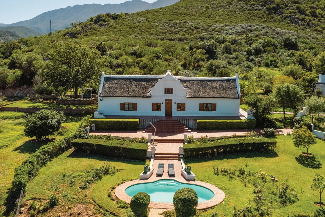 Perfect Hideaways real estate property: Aerial view of a white farmhouse with a thatched roof, nestled in lush green hills. A well-manicured garden and oval pool are in the foreground. Scenic and serene. Cloeten Farm & Guest Cottages, Robertson.