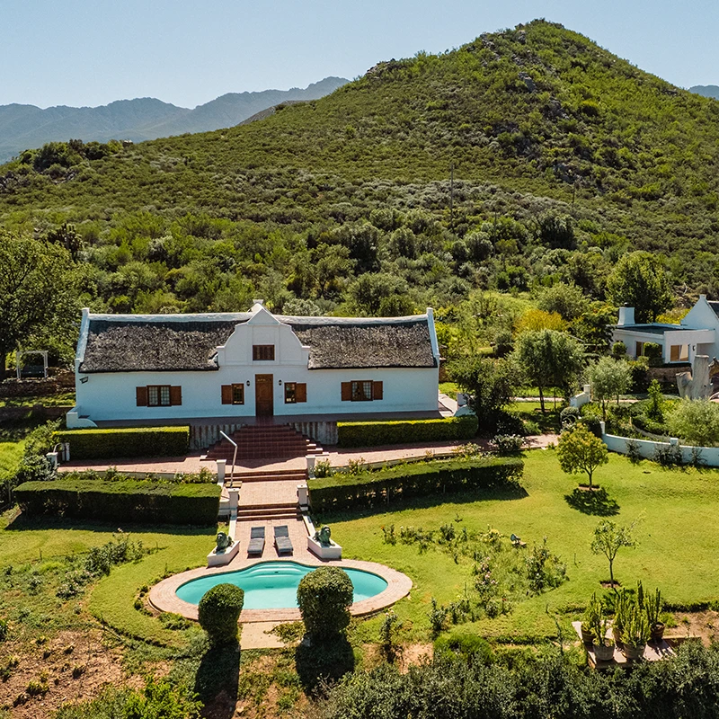 Perfect Hideaways real estate property: Aerial view of a white farmhouse with a thatched roof, nestled in lush green hills. A well-manicured garden and oval pool are in the foreground. Scenic and serene. Cloeten Farm & Guest Cottages, Robertson.
