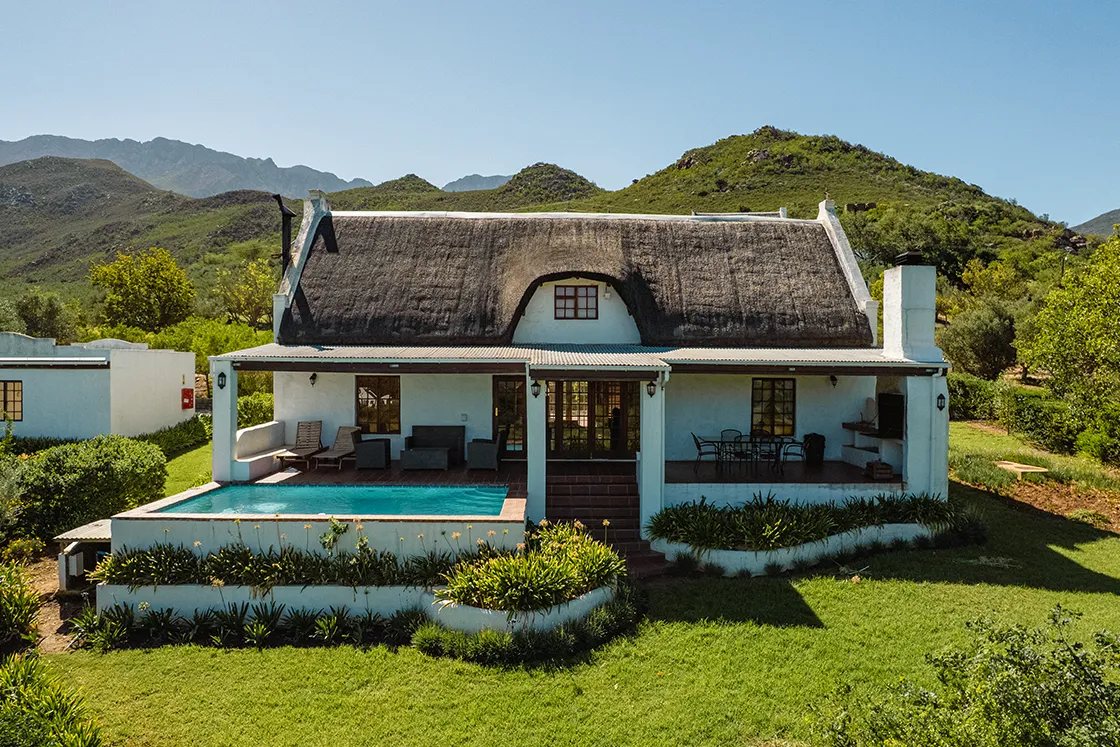 Perfect Hideaways real estate property: Charming cottage with a thatched roof and pool set in lush greenery against a backdrop of rolling hills and blue sky, evoking a tranquil rural vibe. Cloeten Farm & Guest Cottages, Robertson.