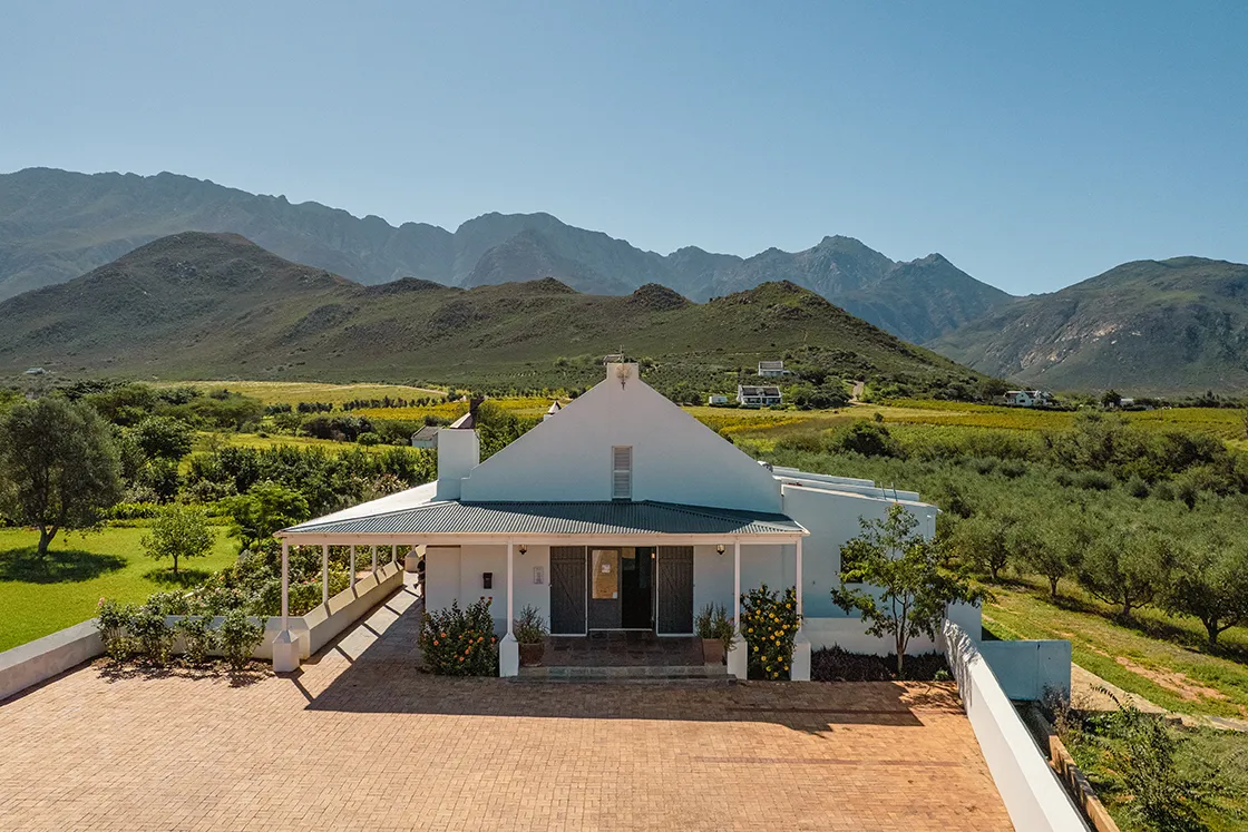 Perfect Hideaways real estate property: Rural farmhouse with a white facade and green roof on a sunny day, set against lush greenery and distant mountains under a clear blue sky. Cloeten Farm & Guest Cottages, Robertson.