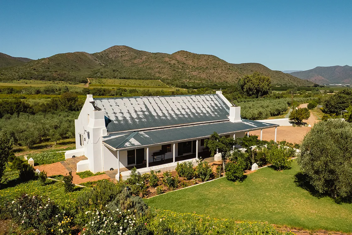 Perfect Hideaways real estate property: A charming white farmhouse with a gray roof sits amid lush greenery, surrounded by mountains under a clear blue sky, conveying a serene rural atmosphere. Cloeten Farm & Guest Cottages, Robertson.
