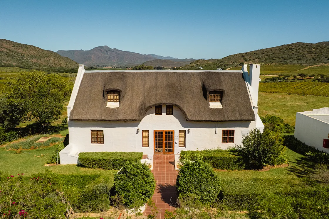 Perfect Hideaways real estate property: A charming white cottage with a thatched roof, set against rolling green hills and mountains. A brick path leads to the wooden front door. Cloeten Farm & Guest Cottages, Robertson.