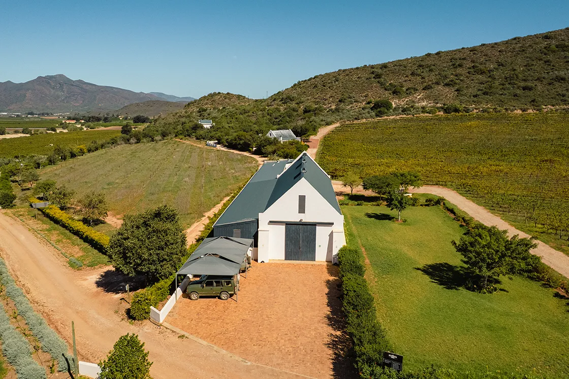 Perfect Hideaways real estate property: Aerial view of a white-roofed building with a parked car under a canopy. It's surrounded by lush greenery, vineyards, and hills under a clear blue sky. Cloeten Farm & Guest Cottages, Robertson.