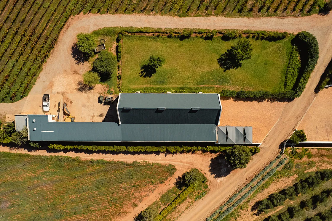 Perfect Hideaways real estate property: Aerial view of a long, rectangular building with a dark roof, surrounded by vineyards and two green lawns. Vehicles and equipment are seen nearby. Cloeten Farm & Guest Cottages, Robertson.