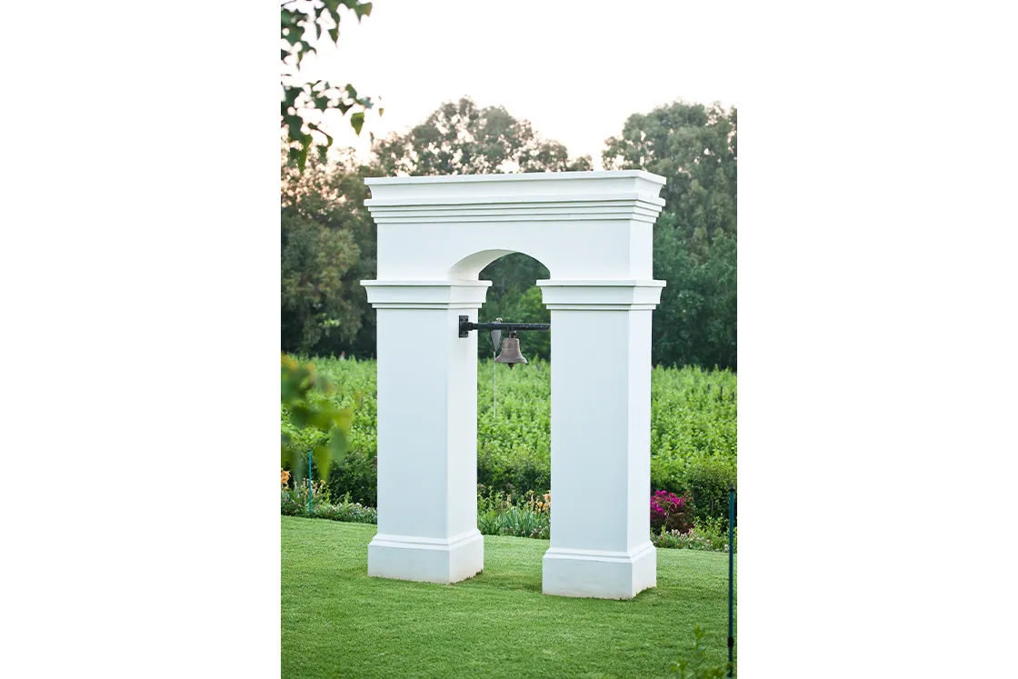 Perfect Hideaways real estate property: White classical garden arch with columns and hanging bell on manicured lawn, lush green vineyard landscape and trees in background, outdoor garden feature, elegant architectural structure. Cloeten Farm & Guest Cottages, Robertson.