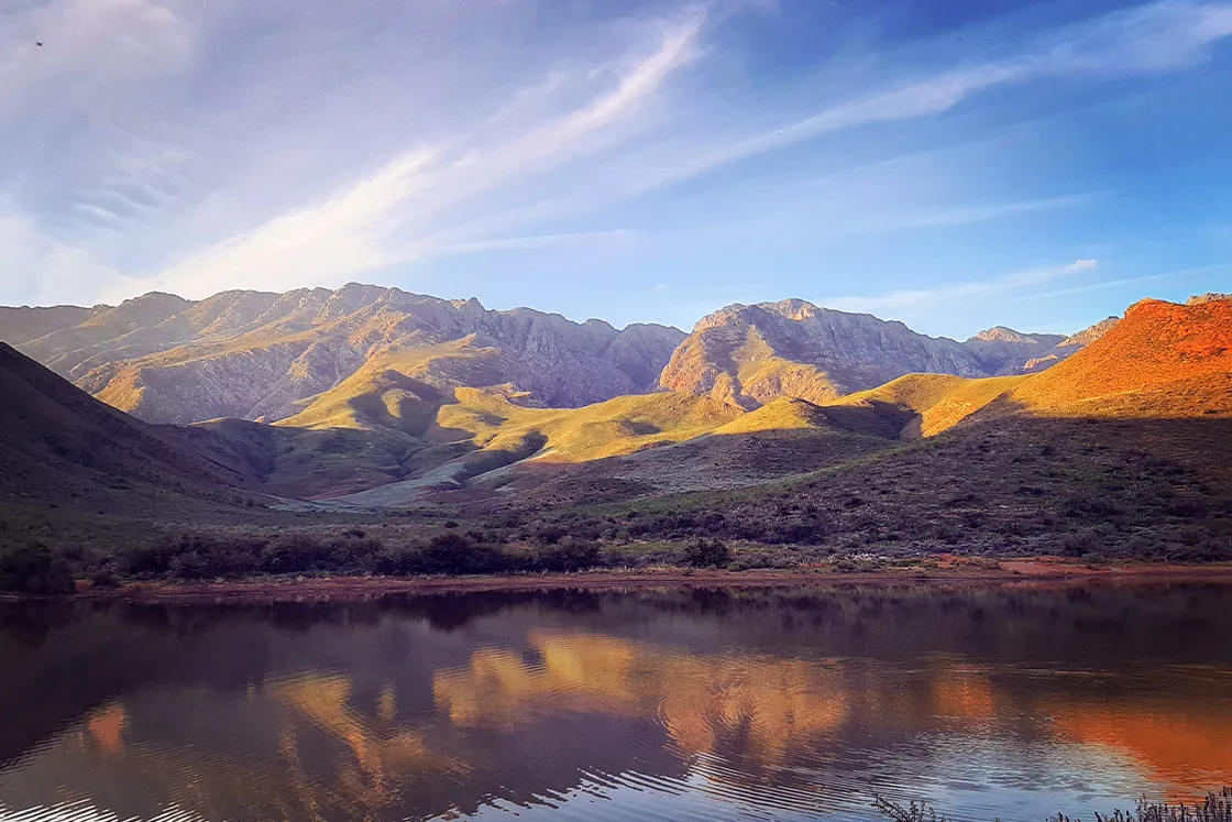 Perfect Hideaways real estate property: Sunlit mountain range reflected in calm lake at golden hour, dramatic shadows on rugged hills, blue sky with wispy clouds, scenic landscape photography, tranquil nature view. Cloeten Farm & Guest Cottages, Robertson.