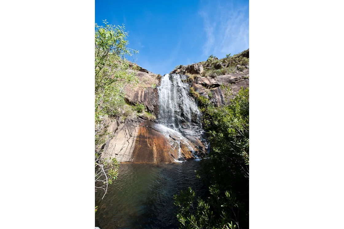 Perfect Hideaways real estate property: Natural waterfall cascading over rocky cliff into clear pool, surrounded by green shrubs and trees under bright blue sky, serene outdoor hiking destination, scenic waterfall landscape. Cloeten Farm & Guest Cottages, Robertson.