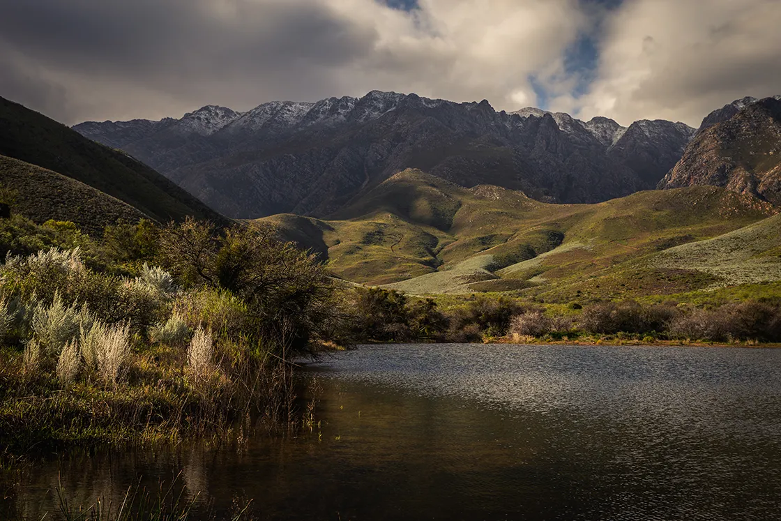 Perfect Hideaways real estate property: Mountain landscape with a tranquil lake in the foreground, surrounded by lush greenery. Snow-capped peaks and dramatic clouds create a serene, majestic scene. Cloeten Farm & Guest Cottages, Robertson.