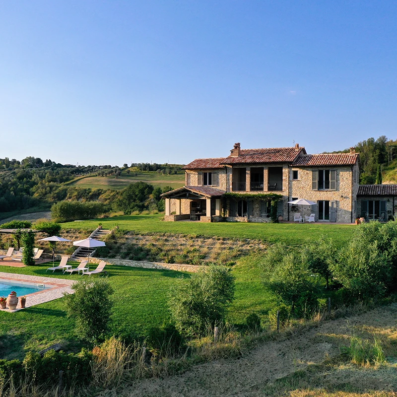 Perfect Hideaways real estate property: Stone villa with red-tiled roof surrounded by lush greenery and rolling hills under a clear blue sky. A pool with loungers sits in the foreground. Casa dell’Oliveto, Narni.