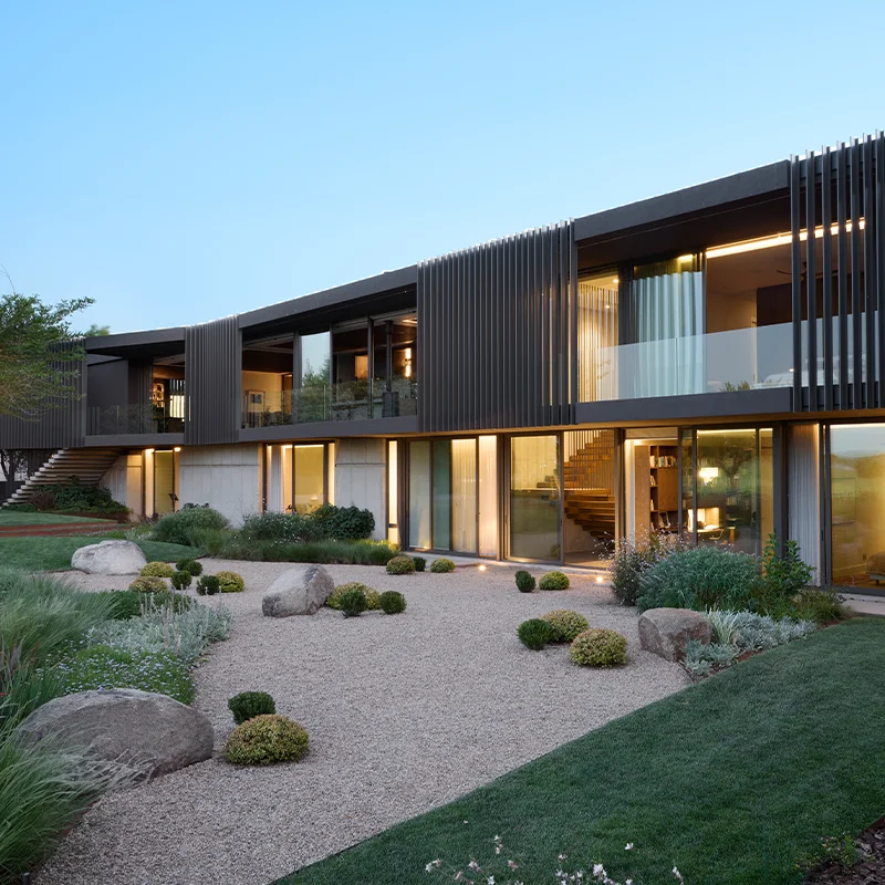 Perfect Hideaways real estate property: Modern two-story house with sleek, dark wood slats, large windows, and landscaped garden. Warm lighting creates an inviting, contemporary feel. Upside-Down House, Kempton Park.