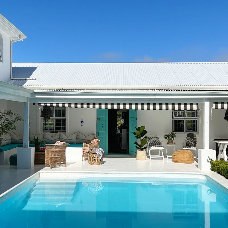 Perfect Hideaways real estate property: Charming poolside patio with turquoise accents and striped awning. Wicker chairs and lush greenery create a relaxing, tropical ambiance under a clear blue sky. Daria, Plettenberg Bay.